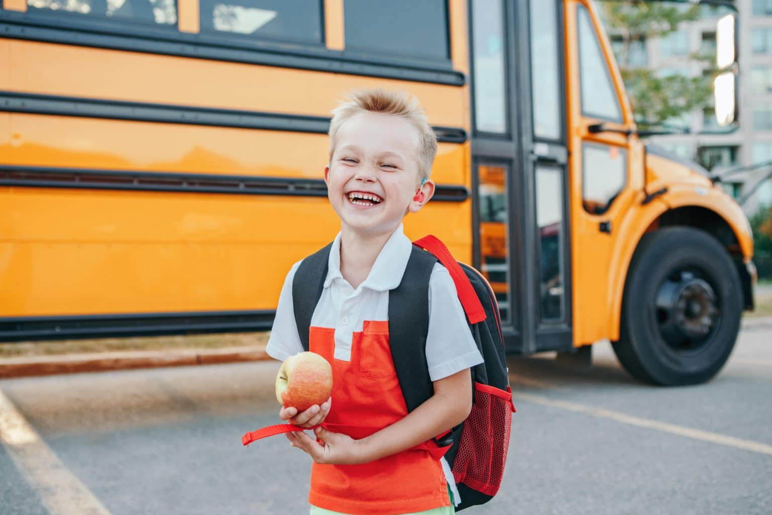 Child wearing pediatric hearing aid Smiling child wearing a backpack and holding an apple. There is a schoolbus parked behind them.